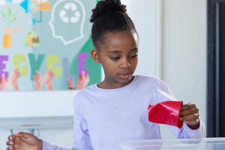 Recycling in school, african american girl placing red paper into recycling bin, focusing on task. environment, sustainability, education, eco-friendly, waste management, responsibilityの写真素材