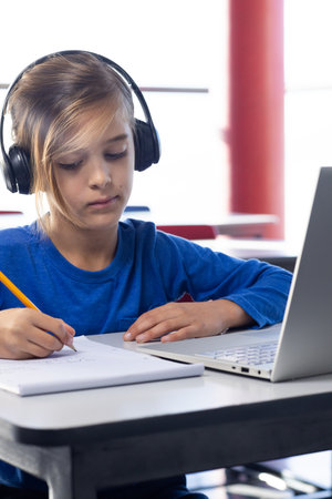 In school, boy wearing headphones writing in notebook and using laptop. Education, learning,...