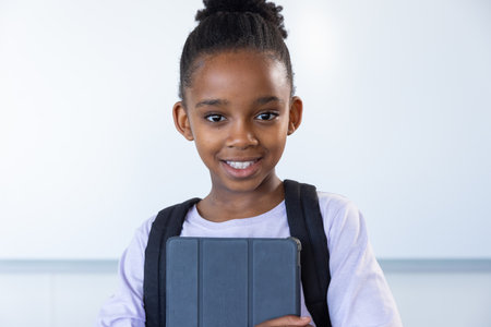 Smiling african american girl holding tablet and wearing backpack in school classroom. Education, student, technology, children, happiness, learningの写真素材