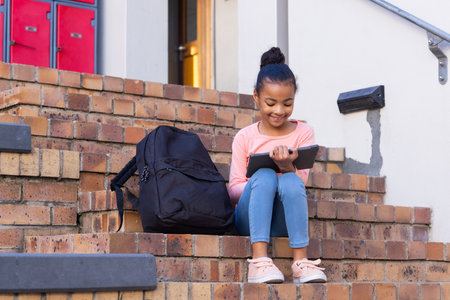 Sitting on school steps, african american girl using tablet with backpack beside her. Education, technology, student, studying, learning, outdoorsの写真素材