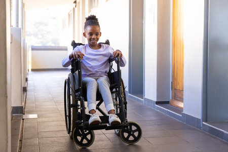 Smiling african american girl in wheelchair moving through school hallway, enjoying her day. Education, disability, inclusivity, independence, happiness, learningの写真素材