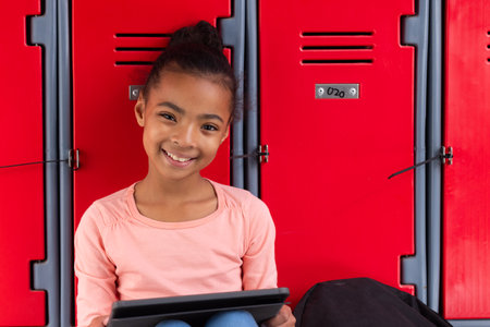 Smiling girl using tablet while sitting in front of red school lockers. Education, back to school, technology, childhood, learning, happinessの写真素材