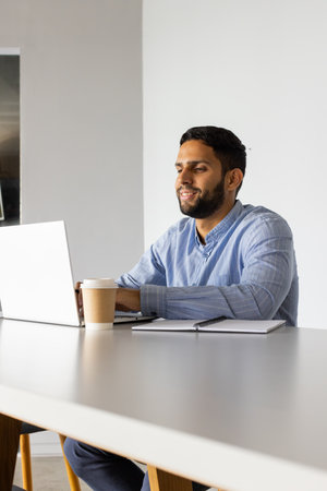 Working on laptop, man smiling with coffee cup and notebook on table. Remote working, home office,...