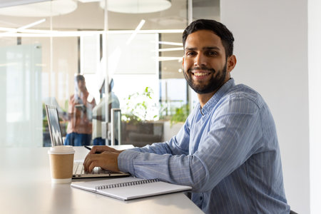 Working on laptop, man smiling with coffee cup and notebook in office. Business, technology,...