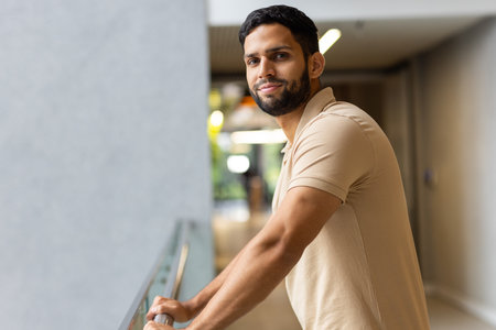 Smiling man leaning on railing in modern office hallway, looking confident, copy space. Business,...