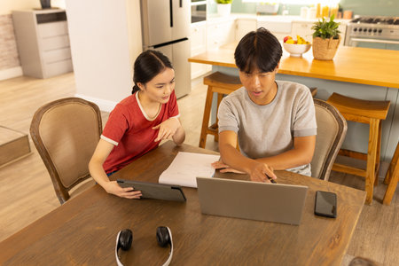 Working from home, asian brother and sister using laptop and tablet at kitchen table. Remote...