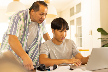 Asian father and teenage son using laptop together at home, focusing on work. family, bonding, technology, parenting, working, togethernessの写真素材