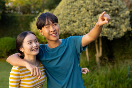 Smiling young asian brother and sister embracing and pointing at something outdoors in park. affection, happiness, love, natureの写真素材