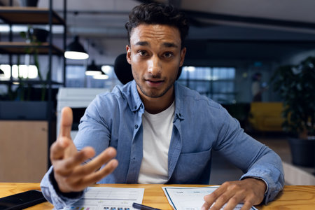 Biracial businessman sitting at desk, making video call, working late at office. Business, overtime,...