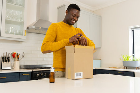 Smiling african american man in kitchen opening package with essential oils, at home. aromatherapy,...