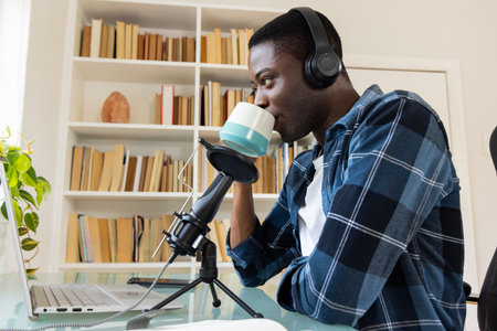 Podcasting at home, African american man drinking coffee and using laptop with headphones. remote...