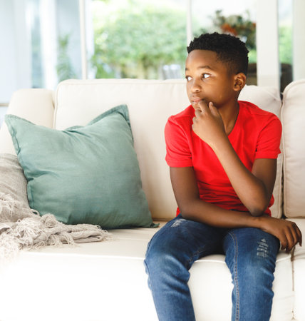 Thoughtful boy in red shirt sitting on cozy sofa at home, copy space. Child, thinking, casual,...