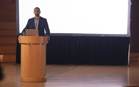 Businessman presenting at conference with laptop on podium, engaging audience, copy space....
