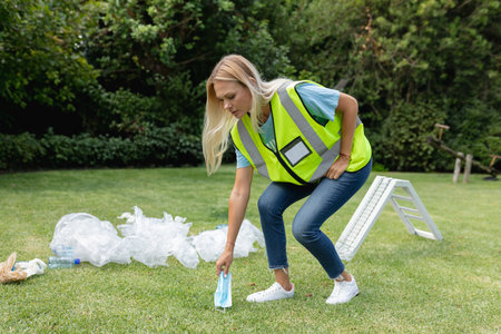Woman in safety vest cleaning up litter in park, promoting environmental care, copy space....