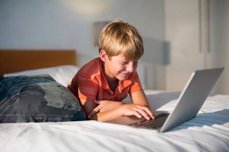 Young boy using laptop on bed at home, smiling and enjoying online activity. Technology, childhood, internet, leisure, wireless, browsingの写真素材
