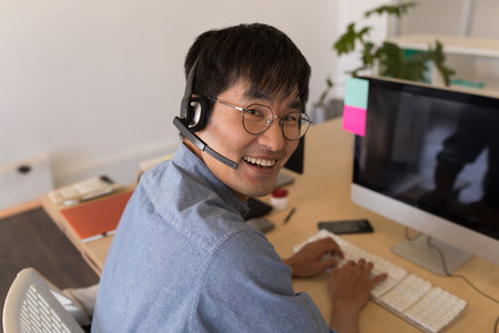 Asian man wearing headset smiling while typing on computer at office desk. Technology,...