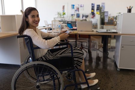 Smiling woman in wheelchair using smartphone at modern office workspace, copy space. technology, communication, business, accessibility, inclusion, digitalの写真素材
