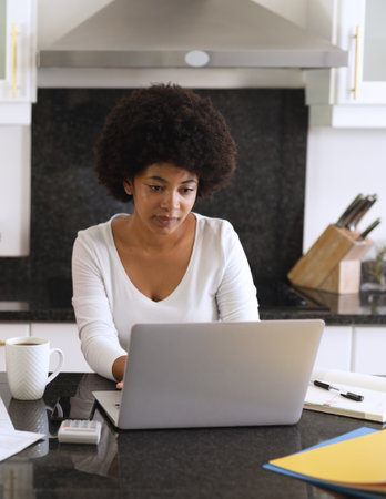 Focused woman working on laptop in modern kitchen with coffee and documents. Remote working, home office, productivity, multitasking, concentration, lifestyleの写真素材