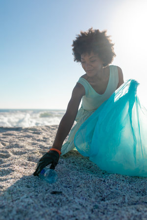 Woman cleaning beach, collecting plastic waste with blue bag under sunny sky. Environment,...