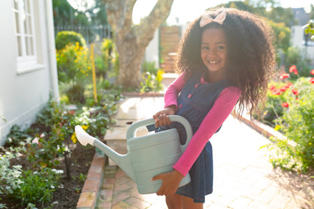 African American girl watering garden with watering can, smiling joyfully outdoors, copy space. Gardening, happiness, child, environmental, hydration, horticultureの写真素材