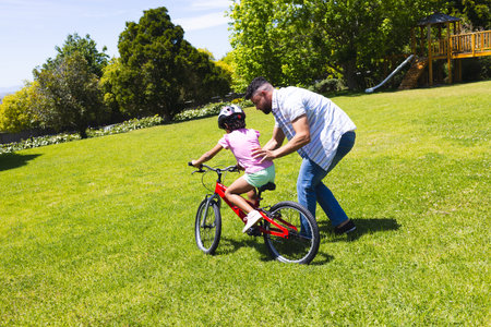 Father teaching daughter to ride bicycle in park, enjoying sunny day together. Family, bonding,...