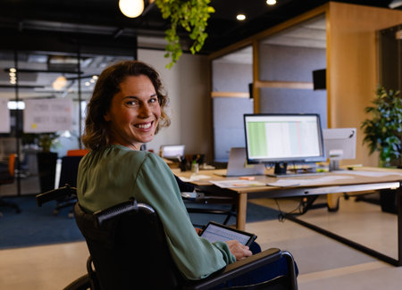 Smiling woman in wheelchair using tablet at modern office desk. Technology, workspace, inclusivity, diversity, digital, professionalの写真素材