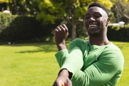 African American man stretching arms in park, smiling and enjoying sunny day, copy space. Outdoors, fitness, exercise, wellness, leisure, happinessの写真素材