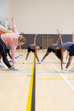 In school gym, physical education class, diverse students stretching with female teacher. fitness,...