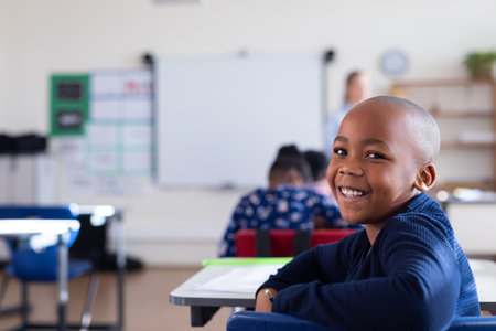 Smiling african american boy sitting at desk in classroom, enjoying school environment. education,...