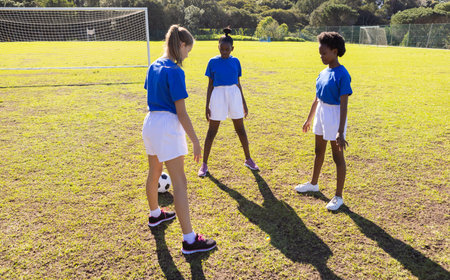 Playing soccer on school field, three diverse girls in sports uniforms practicing skills. teamwork, active, exercise, training, competition, youthの写真素材