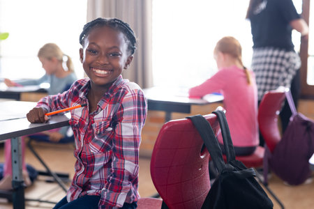 Smiling african american girl holding pencil in classroom with other students studying in school....