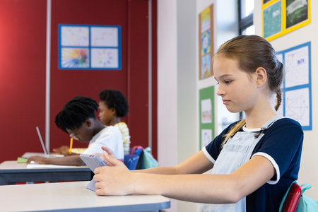 In school, girl using tablet while classmates studying at desks in classroom. Education, technology,...