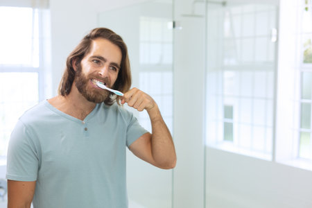 Young man brushing teeth in modern bathroom, with toothbrush and mirror, copy space. Minimalist, contemporary, bright, clean, serene, grooming, oral hygieneの写真素材