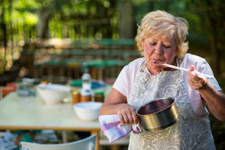 Senior woman in apron tasting sauce in garden patio with saucepan and spoon, copy space. Cooking, sampling, culinary, backyard, alfresco, cookware, rusticの写真素材