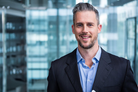 Young adult man standing and smiling in modern office with glass walls, copy space. Businessperson, portrait, corporate, professionalism, confidence, leadership, workspaceの写真素材