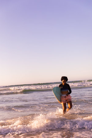 Mid adult African American woman walking in surf on beach holding pastel blue surfboard, copy space. Coastal, shoreline, ocean, watersports, sunlit, golden-hour, serenityの写真素材