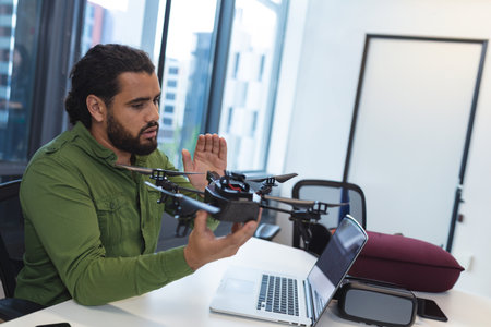 Man inspecting quadcopter drone at modern office desk by window, featuring laptop and VR headset....