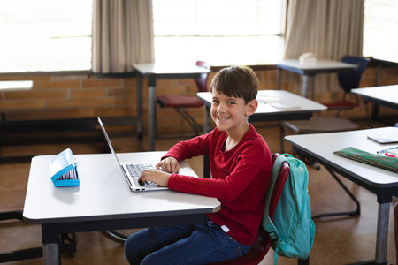 Young boy typing on silver laptop in school classroom, with blue pencil case and teal backpack. Early education, digital learning, educational technology, blended learning, student desk, modern interior, academic environmentの写真素材