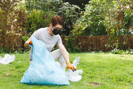 Asian man collecting plastic litter into blue bag on grassy park lawn, wearing orange gloves....