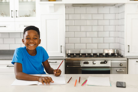 Young African American boy coloring notebook at kitchen island with colored pencils, copy space. Sketching, creativity, education, homework, stationery, countertop, smartphoneの写真素材