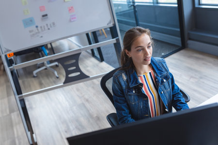 Woman analyzing data on computer monitor while sitting at modern office desk, with whiteboard stand....