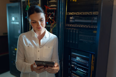 Mid-adult woman using tablet while checking network cables in server room, with blinking LEDs. Data...