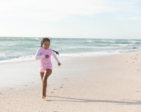bi-racial girl running barefoot along sandy beach shoreline, footprints trailing, copy space. Child, swimwear, pink, seaside, outdoor, active, playfulの写真素材