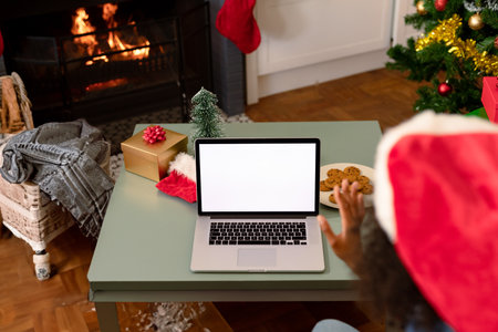 Woman wearing Santa hat sitting at table in living room waving at silver laptop, copy space. Cozy, festive, fireplace, holiday, video call, remote, giftの写真素材