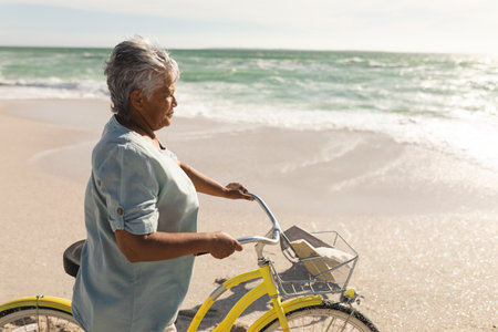 Senior woman pushing yellow bicycle on beach, with basket holding paper bag, book, copy space. Coastal, shoreline, outdoors, sunny, leisure, vacation, retirementの写真素材