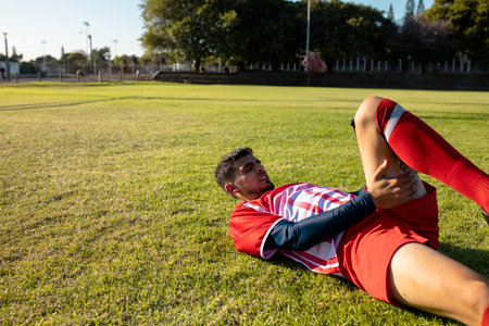 Male soccer player lying on park grass field gripping thigh in discomfort, copy space. Injury, pain, athlete, football, sports, training, rehabilitationの写真素材