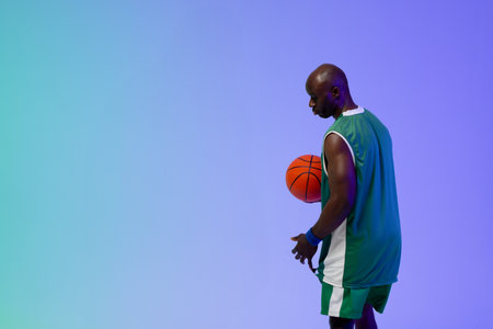 African American basketball player posing in studio, holding basketball in green jersey. Sports ball, sportswear, athletic, male, shorts, wristband, backdropの写真素材