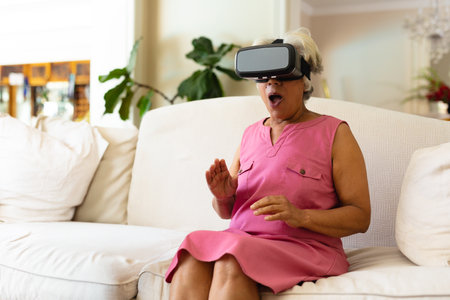 Senior African American woman reacting with surprise wearing VR headset on sofa in living room. Digital, immersive, innovation, entertainment, astonishment, leisure, contemporaryの写真素材