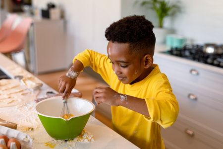 African American boy whisking eggs with metal whisk in green bowl in home kitchen, flour smudges....