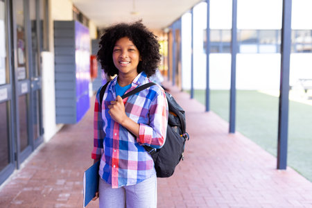 African American boy child standing in school walkway, carrying black backpack, holding blue folder....